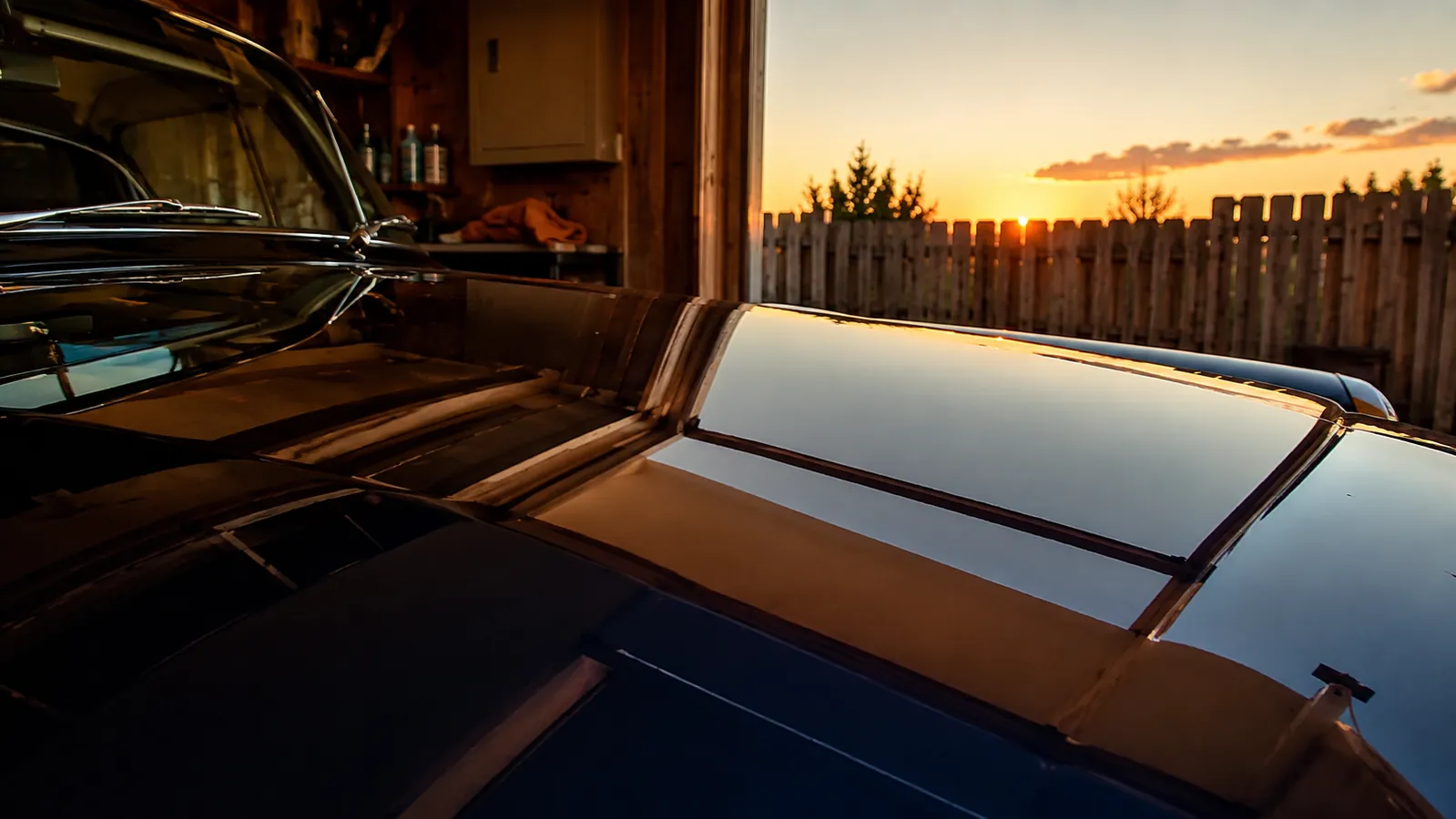 A freshly compounded classic car hood with deep reflections of a garage door and a wooden fence at sunset
