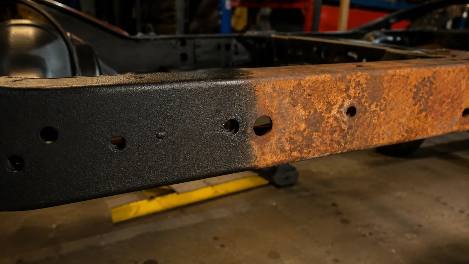 A close-up of a treated metal frame rail with a flat black converted finish beside an untouched patch of orange surface rust on the same metal